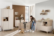 Kinderkamer met een moderne inrichting, inclusief een rechthoekige wandkast aan de muur. Perspectief: zijaanzicht van de kamer met een peuter in een kinderbedje en een vrouw ernaast.