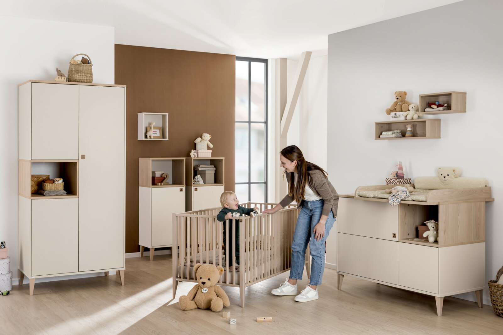Kinderkamer met een moderne inrichting, inclusief een rechthoekige wandkast aan de muur. Perspectief: zijaanzicht van de kamer met een peuter in een kinderbedje en een vrouw ernaast.