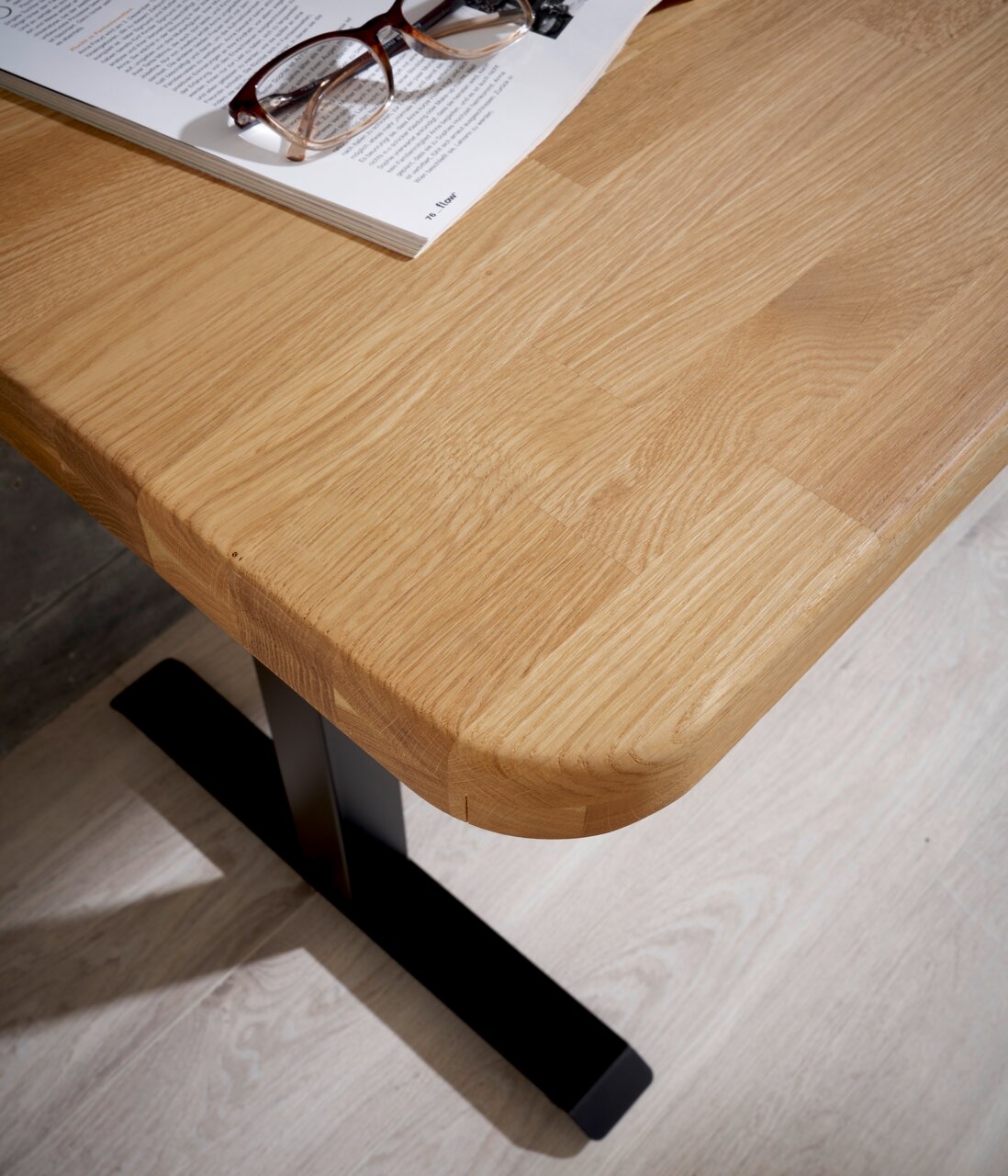 Eckansicht eines E-Schreibtischs mit heller Holzoberfläche und schwarzem Metallfuß, darauf liegen eine Brille und ein aufgeschlagenes Buch.