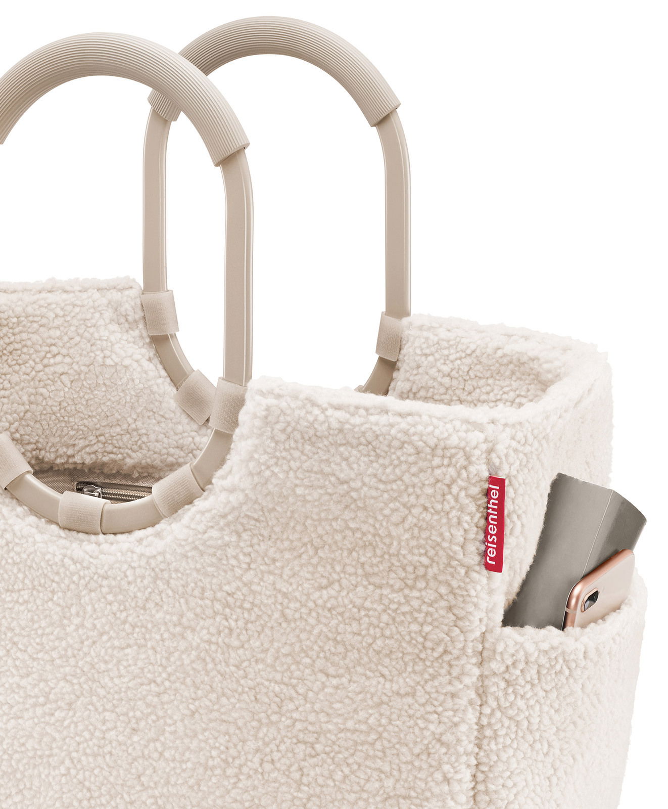 Nahaufnahme eines cremefarbenen, flauschigen Einkaufskorbs mit stabilen Griffen und einer kleinen Außentasche, in der ein Smartphone steckt. Perspektive von oben.