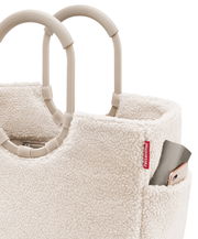 Nahaufnahme eines cremefarbenen, flauschigen Einkaufskorbs mit stabilen Griffen und einer kleinen Außentasche, in der ein Smartphone steckt. Perspektive von oben.