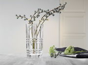 Klare, quadratische Vase aus Glas mit dekorativem Muster, gefüllt mit Zweigen, auf einem Tisch in einem modernen Raum. Perspektive: seitlich.