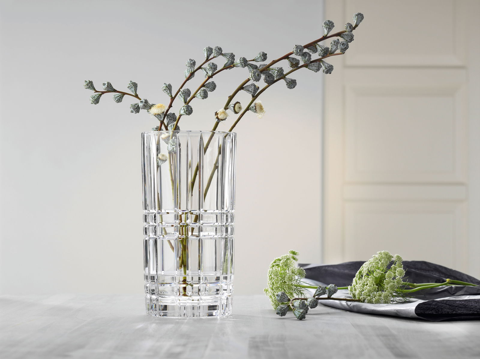 Klare, quadratische Vase aus Glas mit dekorativem Muster, gefüllt mit Zweigen, auf einem Tisch in einem modernen Raum. Perspektive: seitlich.