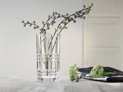 Klare, quadratische Vase aus Glas mit dekorativem Muster, gefüllt mit Zweigen, auf einem Tisch in einem modernen Raum. Perspektive: seitlich.