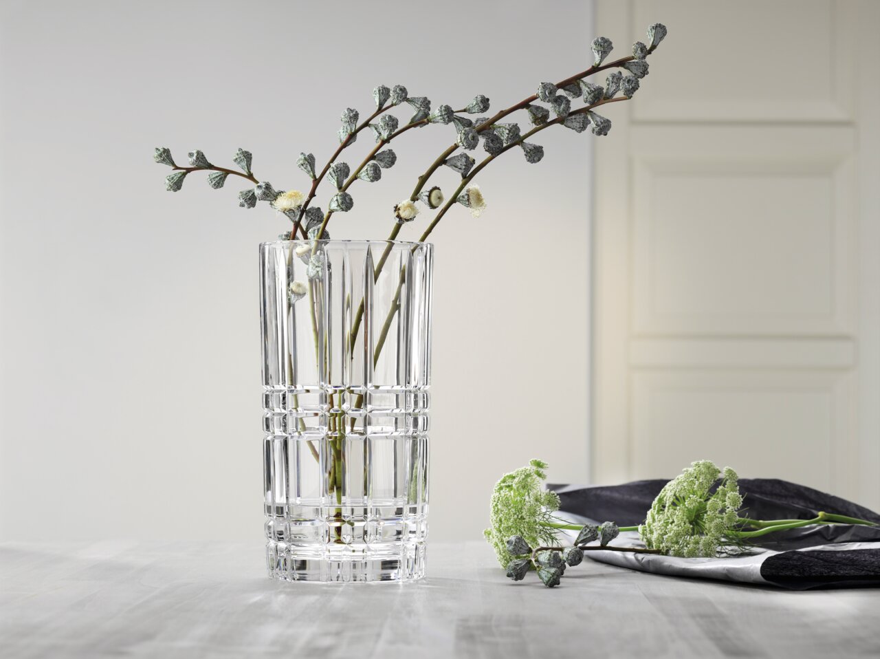 Een heldere, vierkante glazen vaas met een decoratief patroon, gevuld met takjes, op een tafel in een moderne ruimte. Perspectief: zijaanzicht.