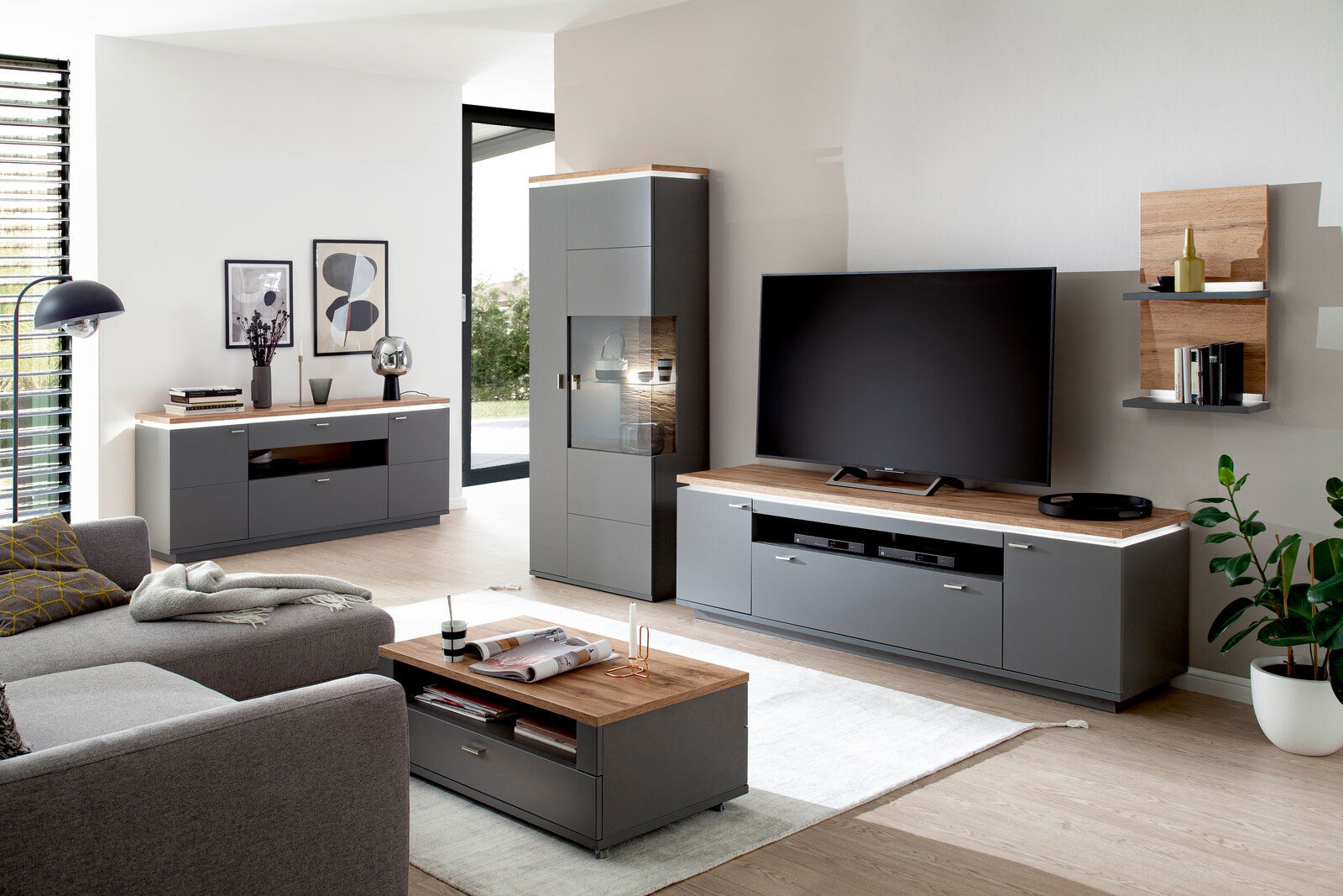 Moderne Wohnzimmermöbel in grauer und holzfarbener Optik, bestehend aus Sideboard, Wandkombination und Couchtisch, aus der Perspektive eines gemütlichen Wohnzimmers.