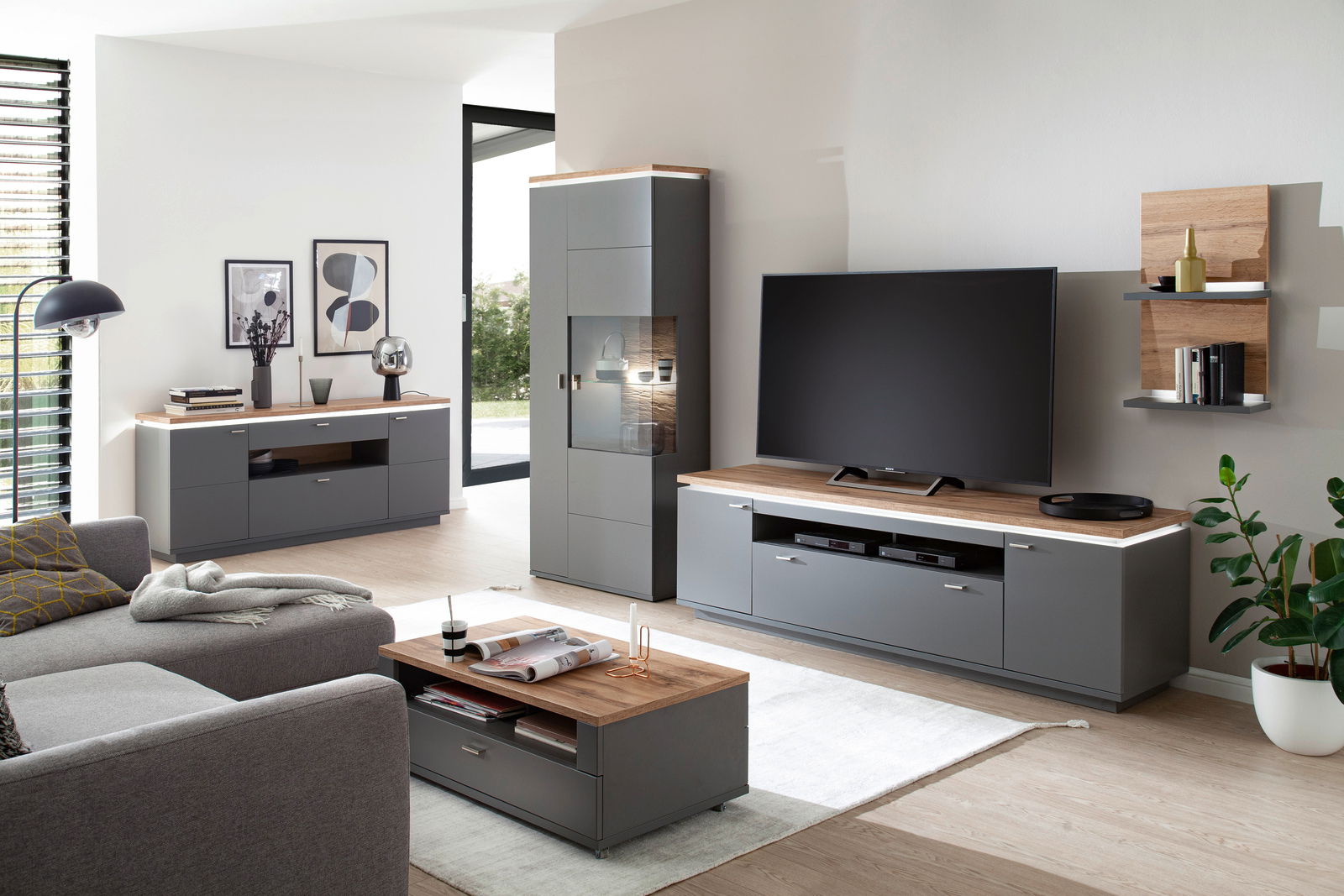 Moderne Wohnzimmermöbel in grauer und holzfarbener Optik, bestehend aus Sideboard, Wandkombination und Couchtisch, aus der Perspektive eines gemütlichen Wohnzimmers.