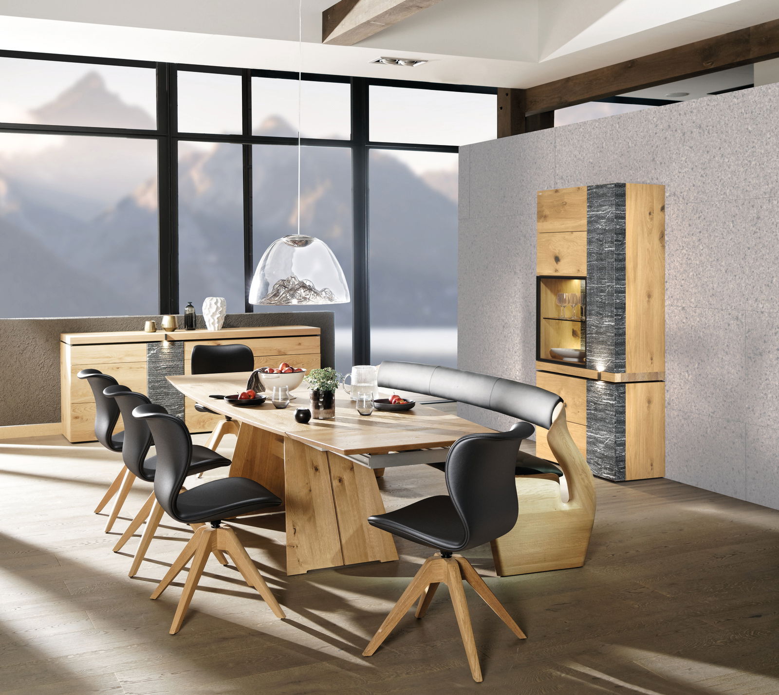 Moderner Essbereich mit Voglauer Esstisch V-ROCK, umgeben von stilvollen Stühlen und einer Eckbank, im Hintergrund ein Sideboard und Highboard aus Holz, mit Bergblick durch große Fenster, Perspektive von schräg vorne.