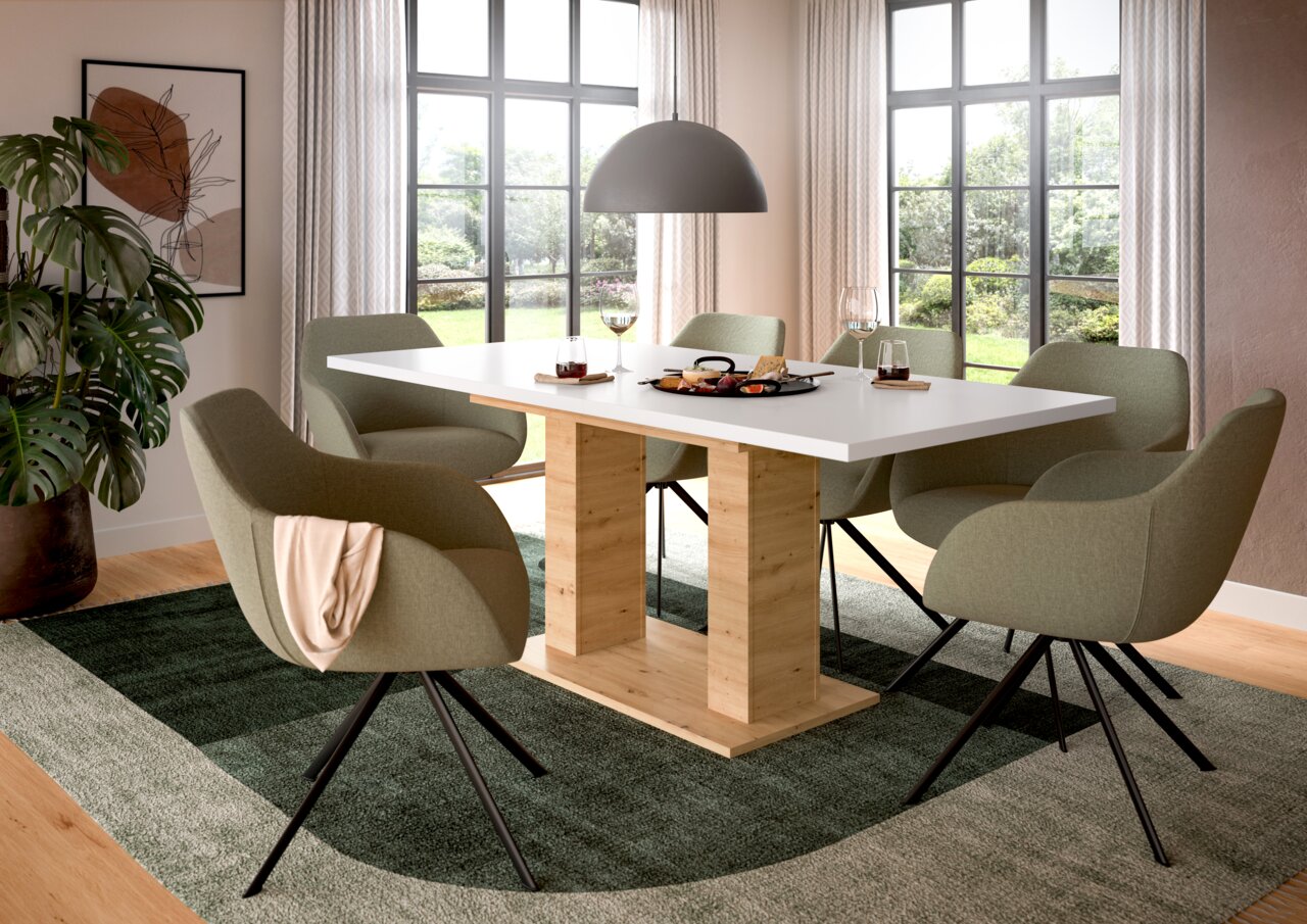 Moderner Esstisch mit weißer Tischplatte und Holzgestell, umgeben von grünen Stühlen in einem hellen Esszimmer mit großen Fenstern, aus seitlicher Perspektive.