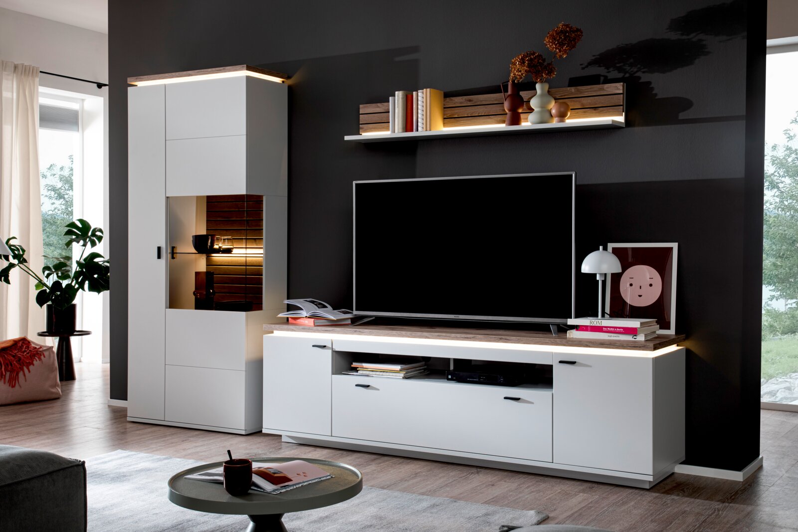 Moderne Wohnwand in Weiß mit Holzoptik-Akzenten, TV-Lowboard, Vitrinenschrank mit Beleuchtung und Wandregal mit LED-Licht, frontal aus leicht schräger Perspektive im Wohnzimmer fotografiert