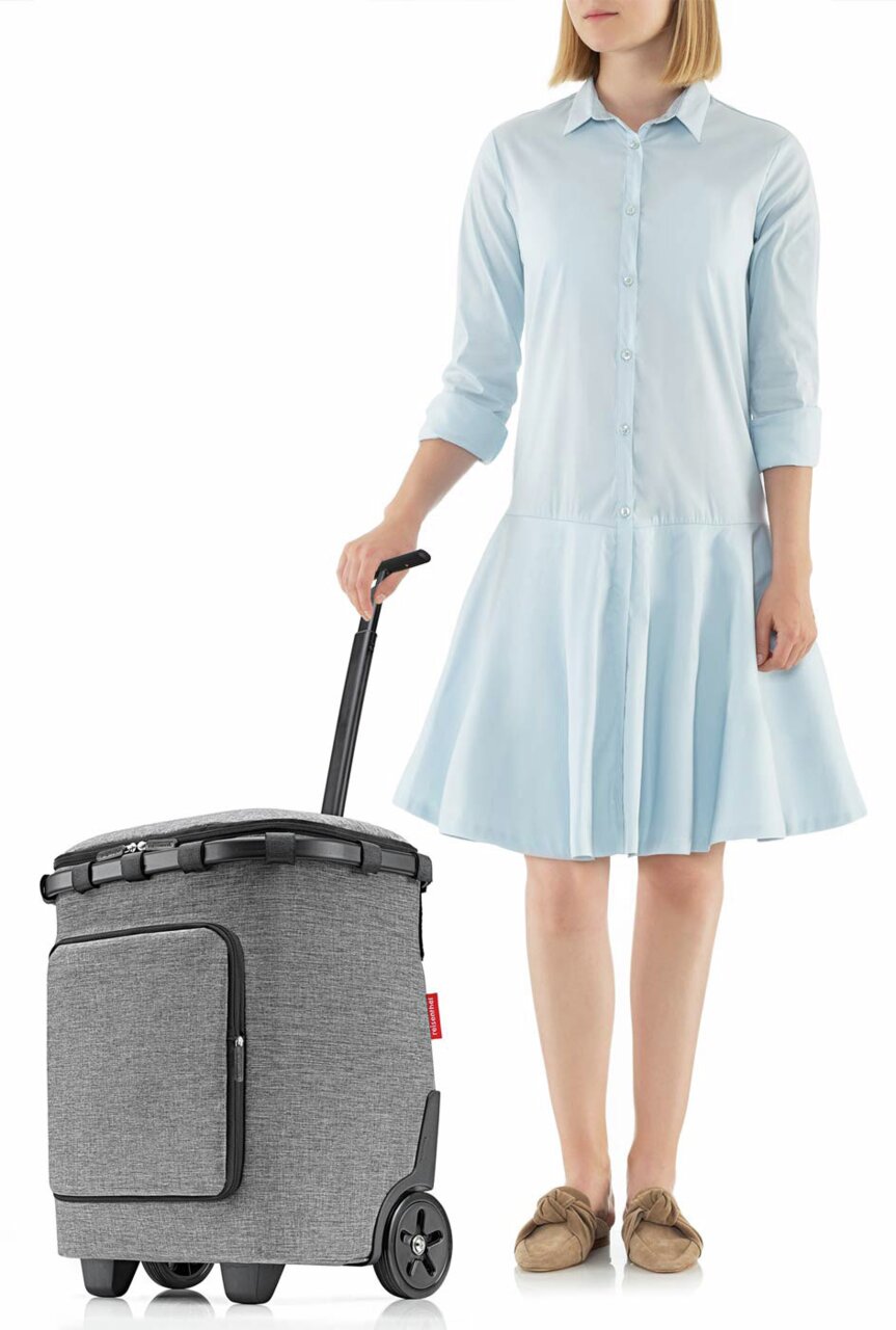 Frau in hellblauem Kleid zieht einen grauen Carrycruiser Plus mit Teleskopgriff und Rädern, seitliche Perspektive.