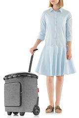 Frau in hellblauem Kleid zieht einen grauen Carrycruiser Plus mit Teleskopgriff und Rädern, seitliche Perspektive.