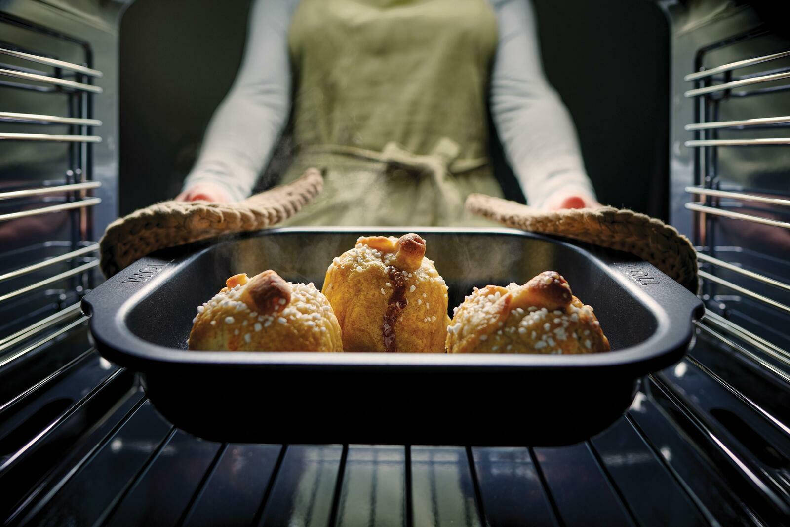 Eine Person nimmt eine Bratreine mit frisch gebackenen Brötchen aus dem Ofen. Die Perspektive ist von vorne auf die Bratreine gerichtet.