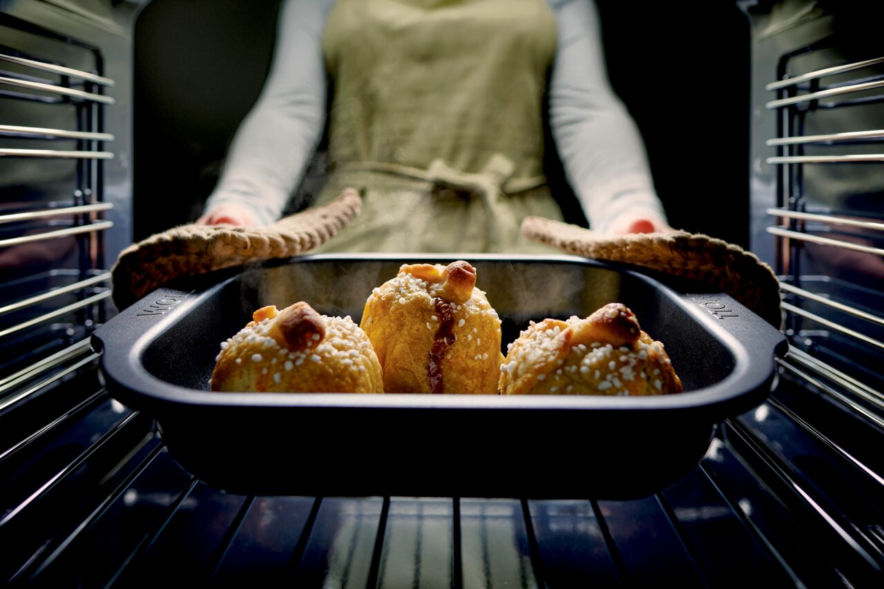 Eine Person nimmt eine Bratreine mit frisch gebackenen Brötchen aus dem Ofen. Die Perspektive ist von vorne auf die Bratreine gerichtet.