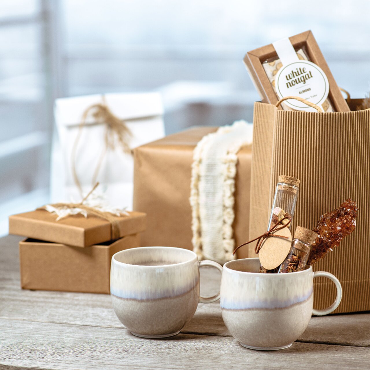 Like by Villeroy & Boch Kaffeebecher PERLEMOR SAND Zwei Keramikbecher mit Henkel in natürlichem Design, umgeben von Geschenkverpackungen, aus seitlicher Perspektive fotografiert.