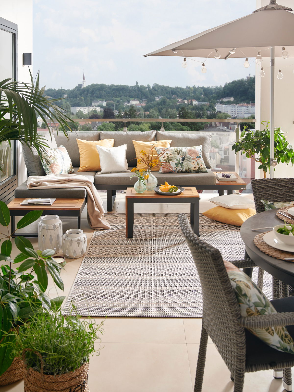 Moderne Gartenlounge mit grauem Sofa, bunten Kissen und einem Tisch auf einer Terrasse mit Blick auf eine grüne Landschaft, aus der Perspektive von vorne.