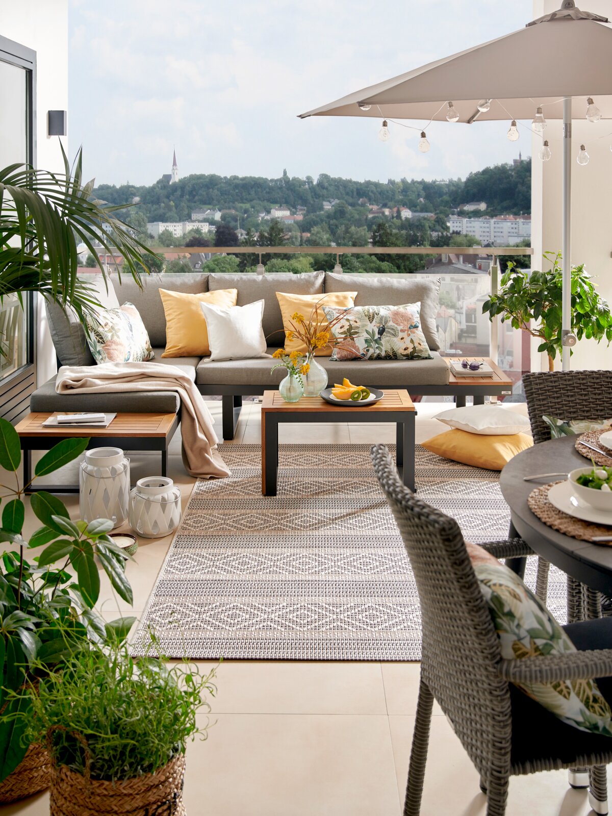 Moderne tuinlounge met een grijze bank, kleurrijke kussens en een tafel op een terras met uitzicht op een groen landschap, gezien van voren.
