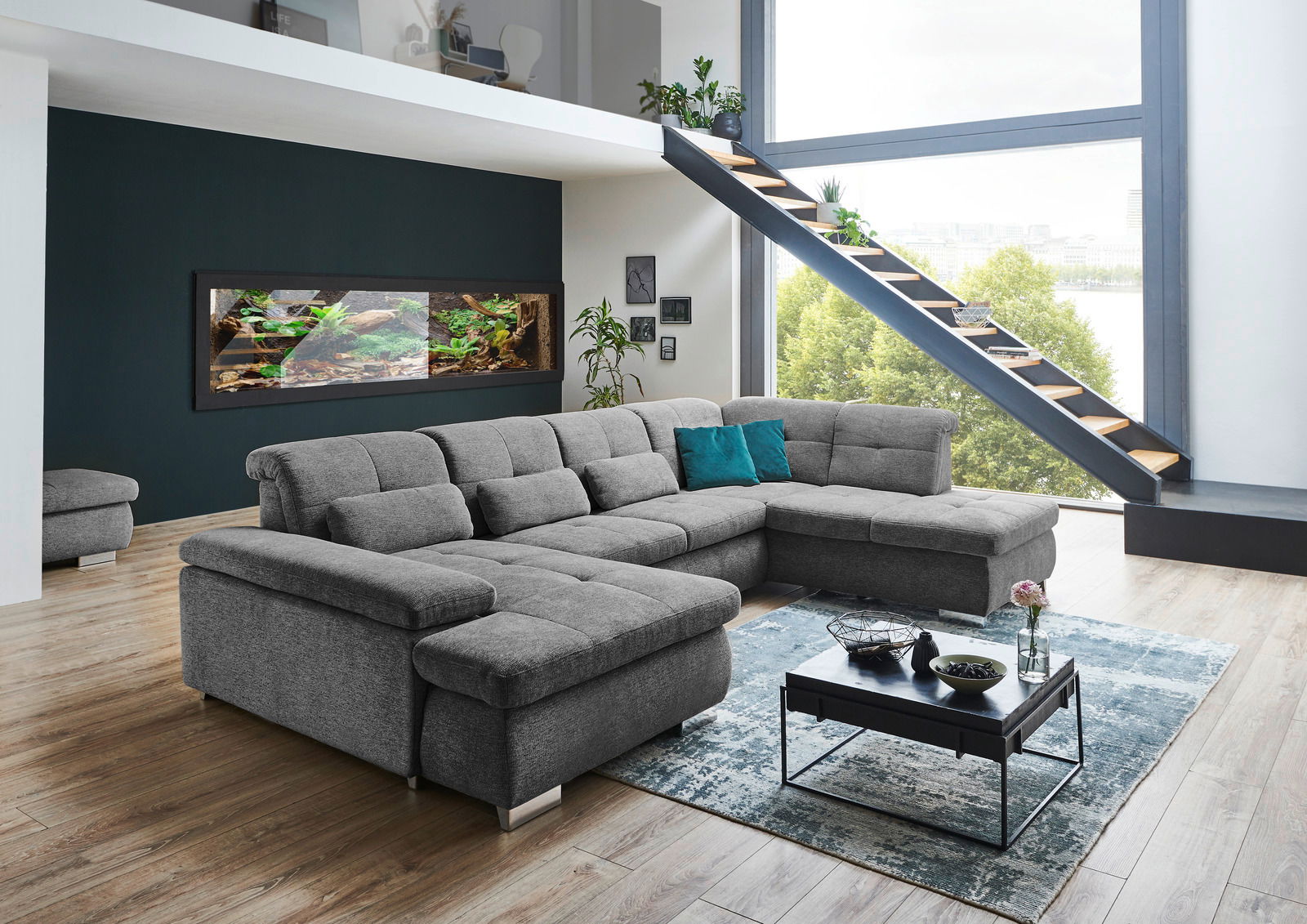 Graue Polsterrundecke Beldomo Style Wohnlandschaft BOCHUM in einem modernen Wohnzimmer mit großen Fenstern und Treppe, seitliche Perspektive.