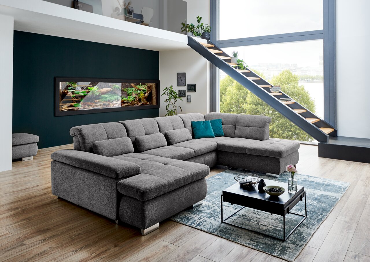 Graue Polsterrundecke Beldomo Style Wohnlandschaft BOCHUM in einem modernen Wohnzimmer mit großen Fenstern und Treppe, seitliche Perspektive.