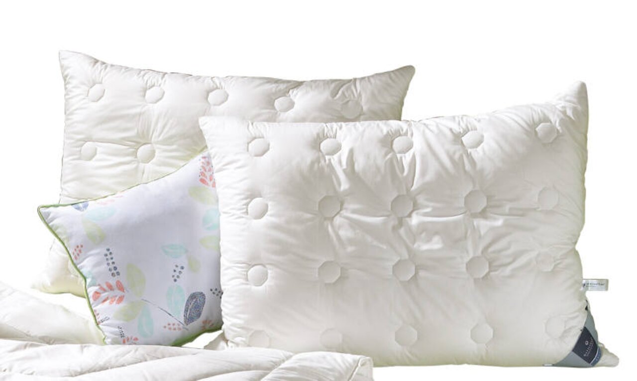 Zwei weiße, gesteppte Kopfkissen der Serie Concerto von billerbeck, frontal abgebildet, mit einem dekorativen Kissen im Hintergrund.