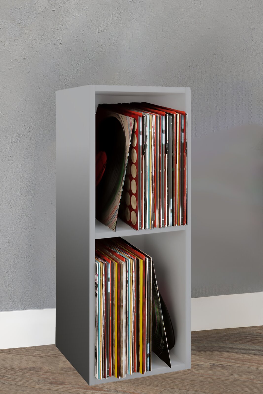 Schmales, weißes Schallplatten-Regal mit zwei Fächern, gefüllt mit Vinylplatten, in leicht seitlicher Frontansicht vor grauer Wand auf Holzboden