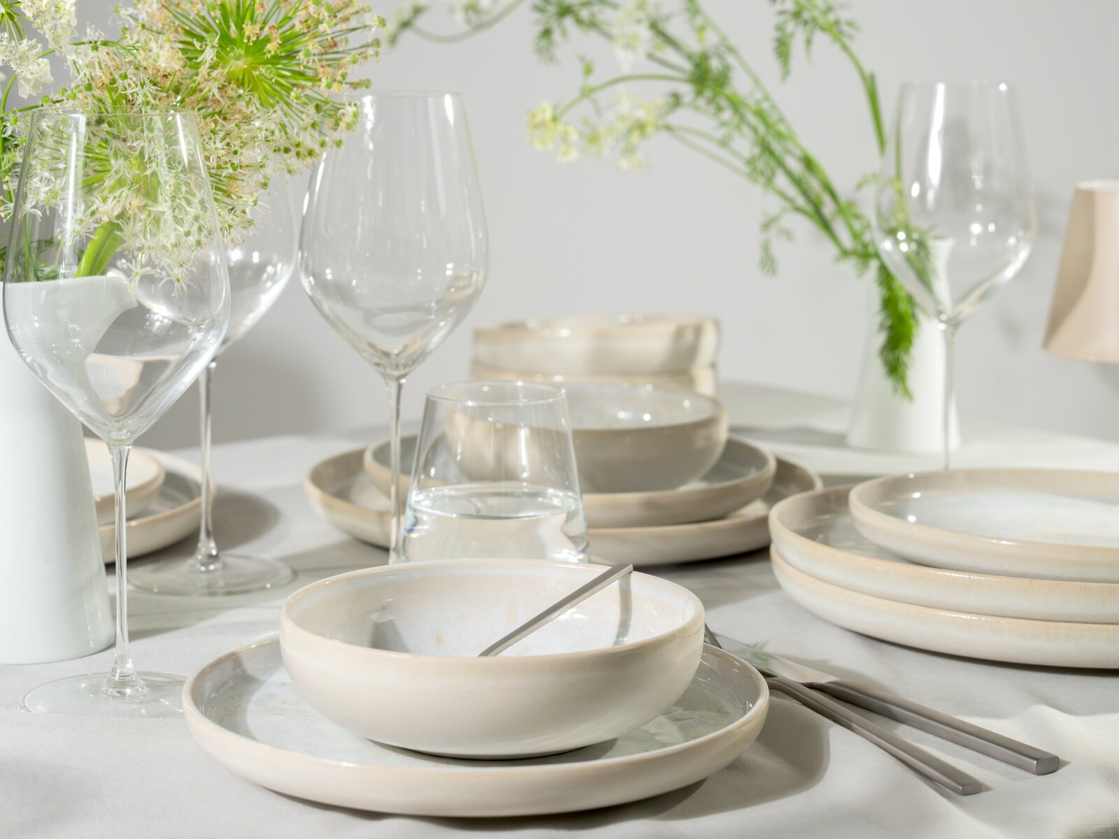 Nahaufnahme aus leicht erhöhter Frontperspektive auf eine elegant gedeckte Tafel mit cremefarbenem Keramikgeschirr, Weingläsern, Besteck und weißen Blumen.