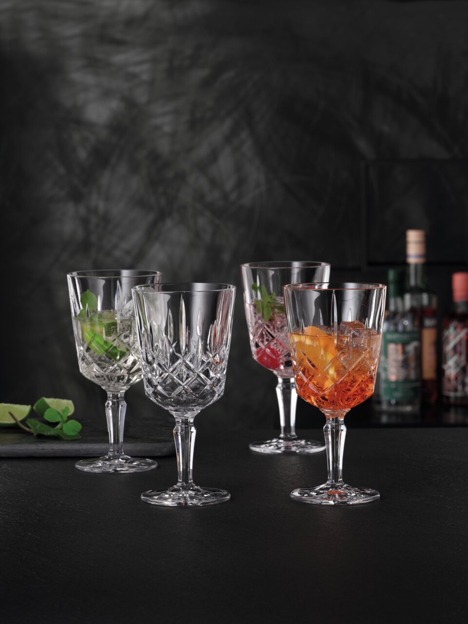 Vier elegante Cocktailgläser Noblesse mit facettiertem Design, gefüllt mit bunten Getränken, auf schwarzem Hintergrund. Frontale Perspektive.