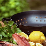 Schwarze BBQ Wokpfanne mit Löchern, umgeben von frischen Zutaten wie Zitrone und Kräutern, aus seitlicher Perspektive fotografiert.