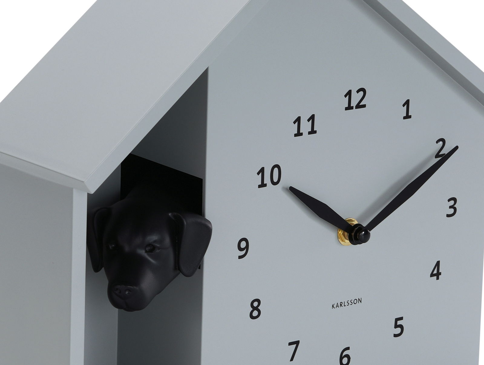 Moderne Wanduhr in Form eines Hauses mit einem schwarzen Hundekopf, der aus einer Öffnung herausschaut. Die Uhr hat ein minimalistisches Design mit schwarzen Zeigern und Zahlen auf einem hellgrauen Hintergrund. Perspektive von schräg vorne.