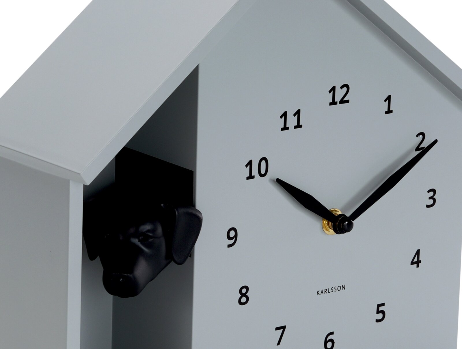 Moderne wandklok in de vorm van een huis met een zwarte hondenkop die uit een opening gluurt. De klok heeft een minimalistisch ontwerp met zwarte wijzers en cijfers op een lichtgrijze achtergrond. Weergave vanuit een schuine hoek van voren.