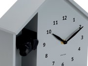 Moderne wandklok in de vorm van een huis met een zwarte hondenkop die uit een opening gluurt. De klok heeft een minimalistisch ontwerp met zwarte wijzers en cijfers op een lichtgrijze achtergrond. Weergave vanuit een schuine hoek van voren.