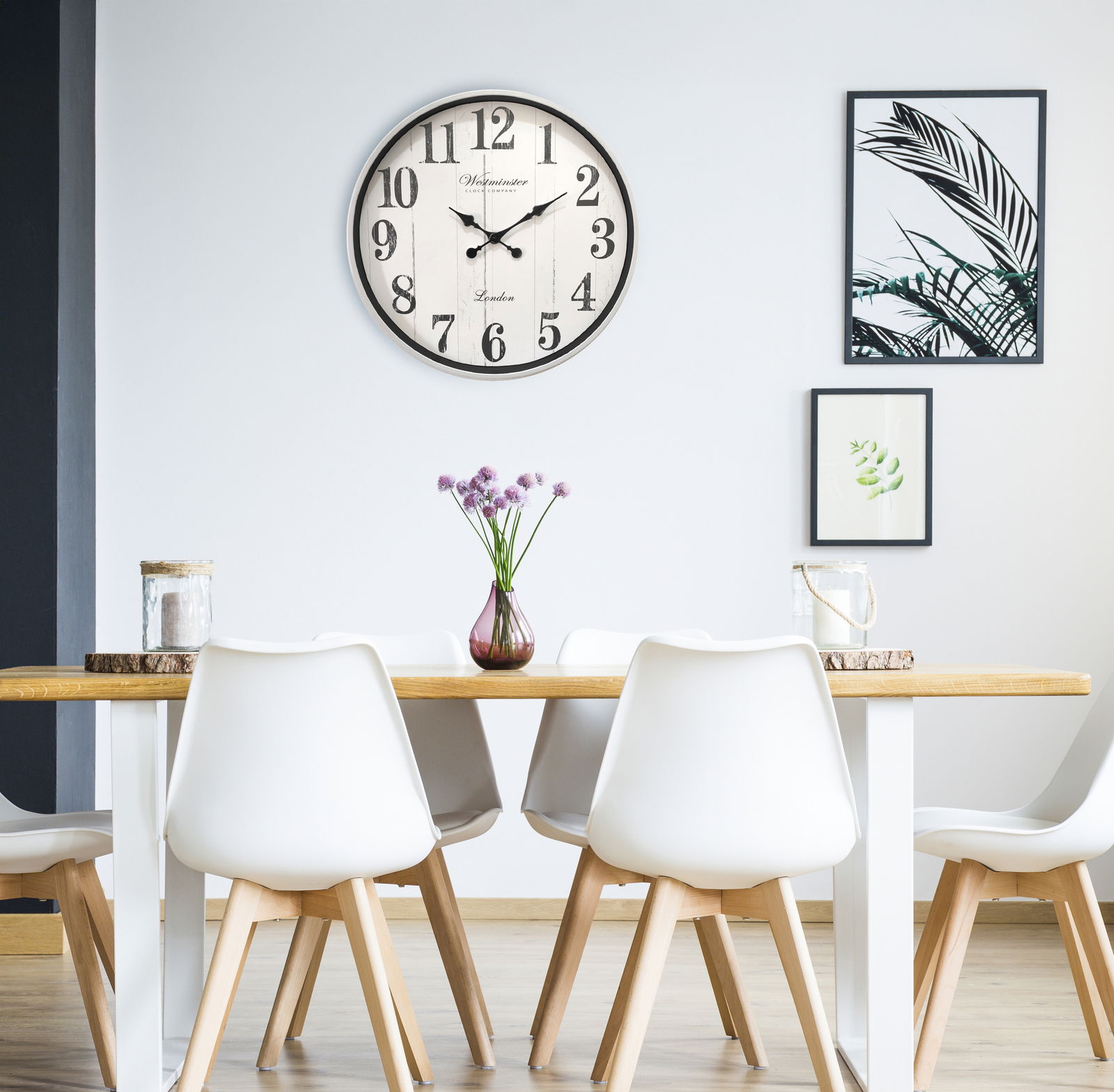 Moderne Wanduhr mit großen schwarzen Ziffern und Zeigern, aufgehängt über einem stilvollen Essbereich mit weißen Stühlen und Holztisch. Perspektive: Frontalansicht.