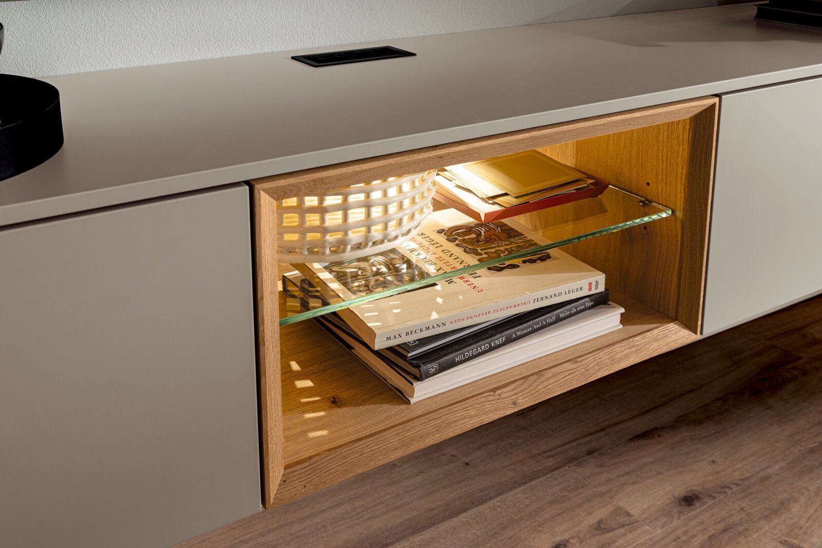 Nahaufnahme einer modernen Wandkombination mit Holz- und Glaselementen, beleuchtetem Regal und dekorativen Büchern, seitliche Perspektive.