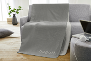 Graue Jacquard-Decke mit 'bugatti'-Schriftzug, drapiert über einem grauen Sofa in einem modernen Wohnzimmer.
