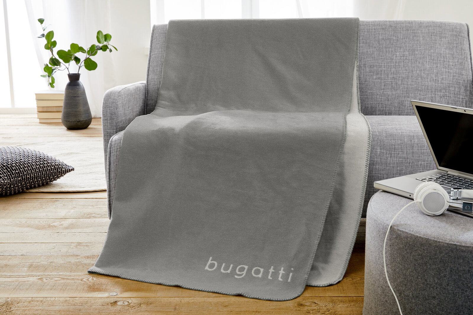 Graue Jacquard-Decke mit 'bugatti'-Schriftzug, drapiert über einem grauen Sofa in einem modernen Wohnzimmer.