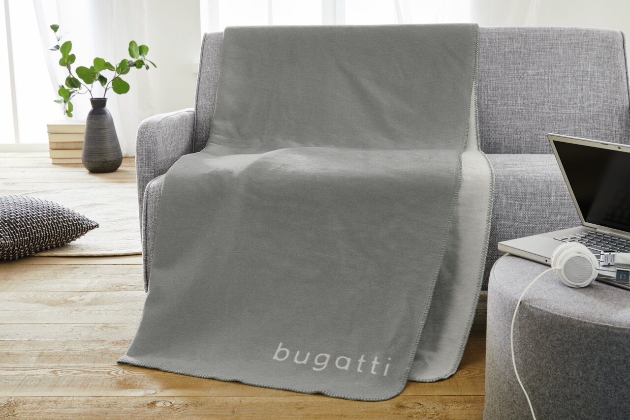 Graue Jacquard-Decke mit 'bugatti'-Schriftzug, drapiert über einem grauen Sofa in einem modernen Wohnzimmer.