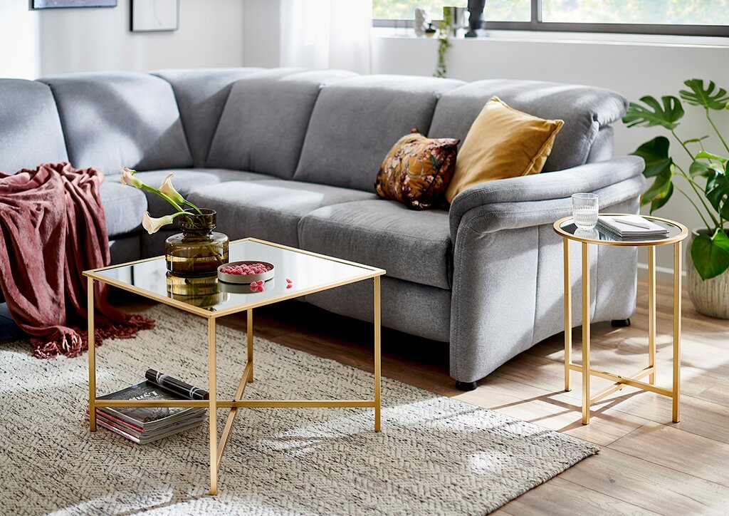 Modernes Wohnzimmer mit grauem Ecksofa, goldfarbenem Couchtisch mit Glasplatte und passendem Beistelltisch; Perspektive: leicht schräg von vorn auf die Sofaecke und die Tische.