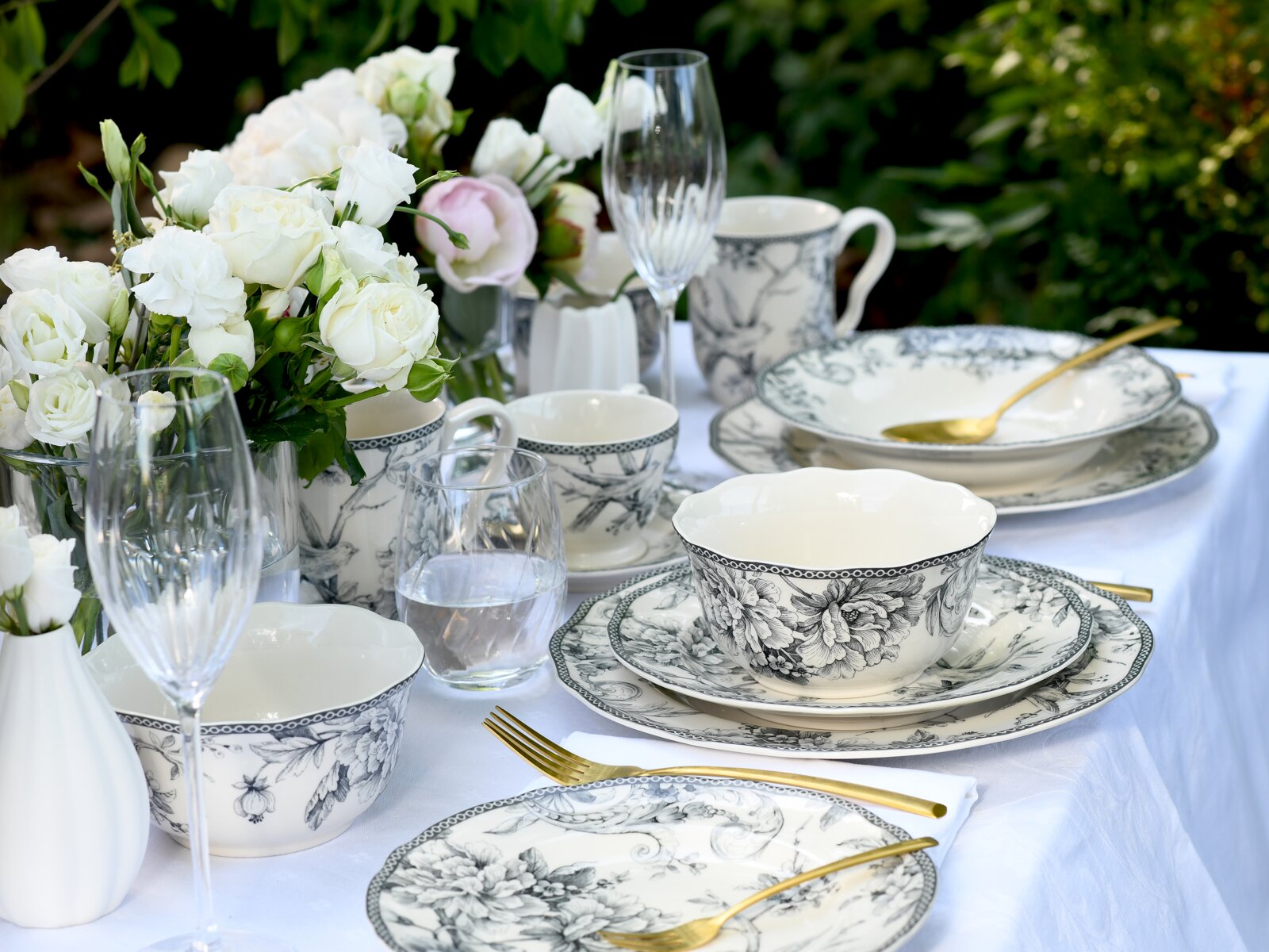 Schwarz-weißes Porzellan-Geschirrset mit floralen Motiven auf gedecktem Outdoor-Tisch, mit goldfarbenem Besteck, Gläsern und weißen Rosen; Perspektive: leicht seitliche Nahaufnahme auf Tischhöhe.