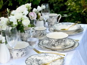 Schwarz-weißes Porzellan-Geschirrset mit floralen Motiven auf gedecktem Outdoor-Tisch, mit goldfarbenem Besteck, Gläsern und weißen Rosen; Perspektive: leicht seitliche Nahaufnahme auf Tischhöhe.