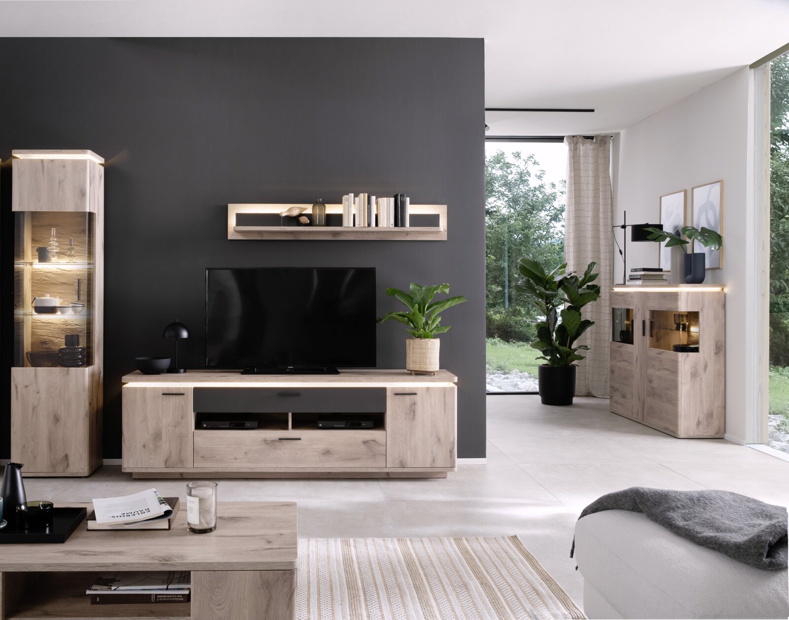 Moderne woonkamerinrichting met meubels in licht hout en antraciet: tv-kast met televisie, wandrek, verlichte vitrinekast en dressoir, gefotografeerd vanuit een vooraanzicht.