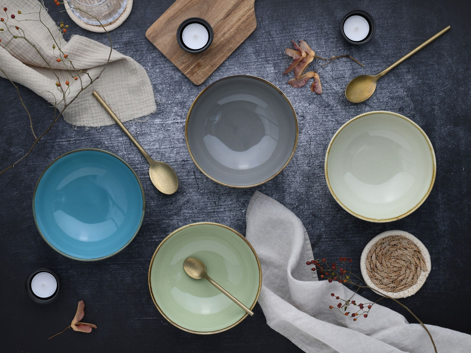 Vogelperspektive auf ein Set aus vier bunten Schalen in Blau, Grau, Grün und Creme, arrangiert mit goldenen Löffeln und dekorativen Elementen auf einem dunklen Untergrund.