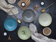 Vogelperspektive auf ein Set aus vier bunten Schalen in Blau, Grau, Grün und Creme, arrangiert mit goldenen Löffeln und dekorativen Elementen auf einem dunklen Untergrund.