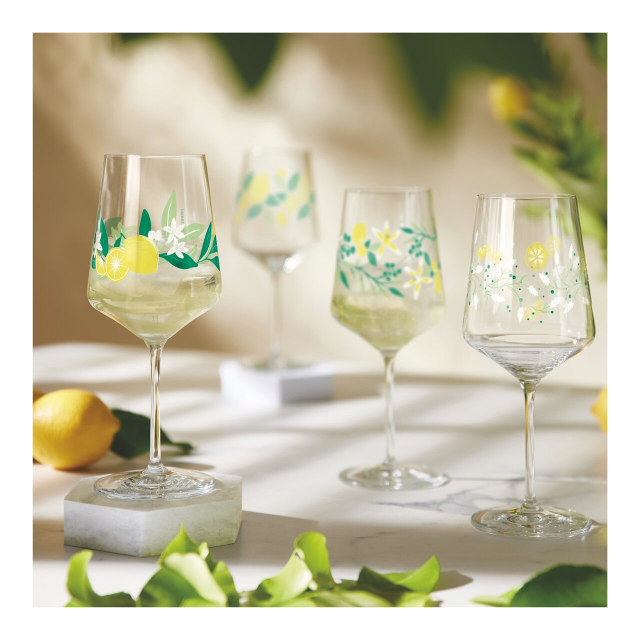 Vier elegante Weingläser mit Zitronen- und Blumenmotiven, auf einem Tisch arrangiert, mit einem frischen und sommerlichen Ambiente.