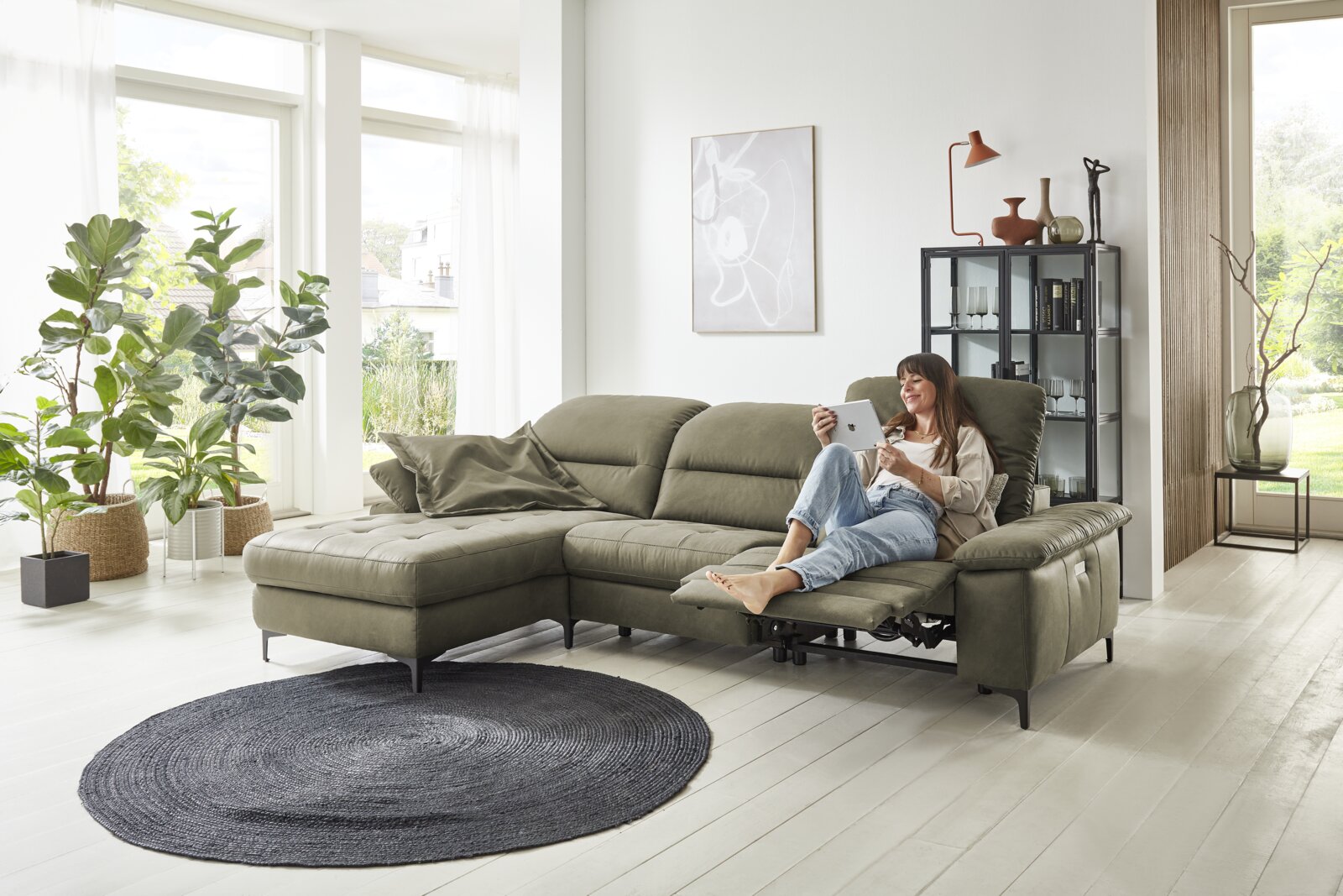 Gemütliche Longchairkombination in einem modernen Wohnzimmer mit großen Fenstern, Pflanzen und einer entspannten Person, die auf dem Sofa sitzt. Perspektive von vorne.