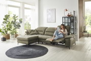 Gemütliche Longchairkombination in einem modernen Wohnzimmer mit großen Fenstern, Pflanzen und einer entspannten Person, die auf dem Sofa sitzt. Perspektive von vorne.