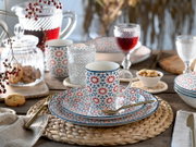 Schräg von vorn fotografiertes Tischgedeck mit weißer Tasse und passendem Teller im rot-blauen Sternmuster, umgeben von Gläsern, Karaffe mit rotem Getränk und kleinen Snacks auf Holztisch.