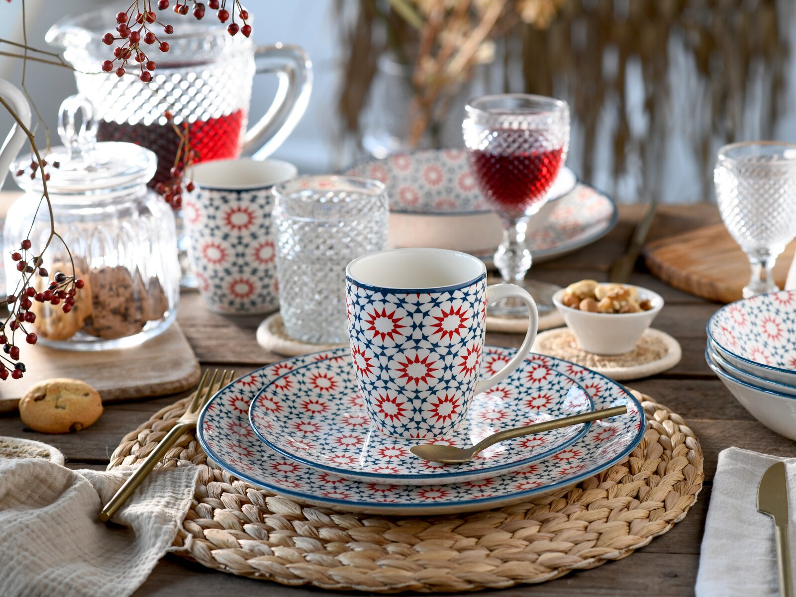 Schräg von vorn fotografiertes Tischgedeck mit weißer Tasse und passendem Teller im rot-blauen Sternmuster, umgeben von Gläsern, Karaffe mit rotem Getränk und kleinen Snacks auf Holztisch.