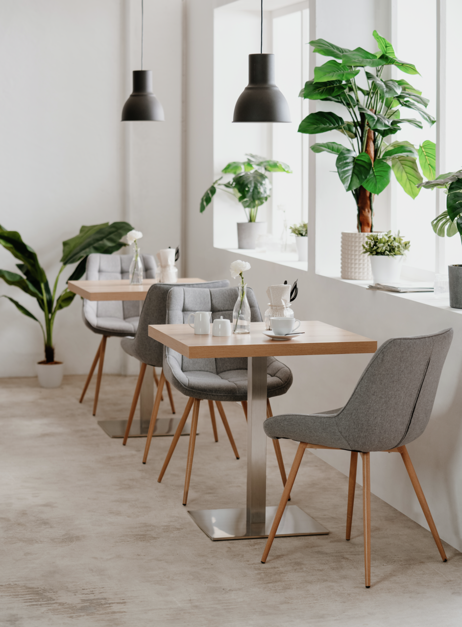 Moderner Esstisch mit quadratischer Holzplatte und Metallfuß, umgeben von grauen Polsterstühlen in einem hellen Raum mit Pflanzen, seitliche Perspektive.