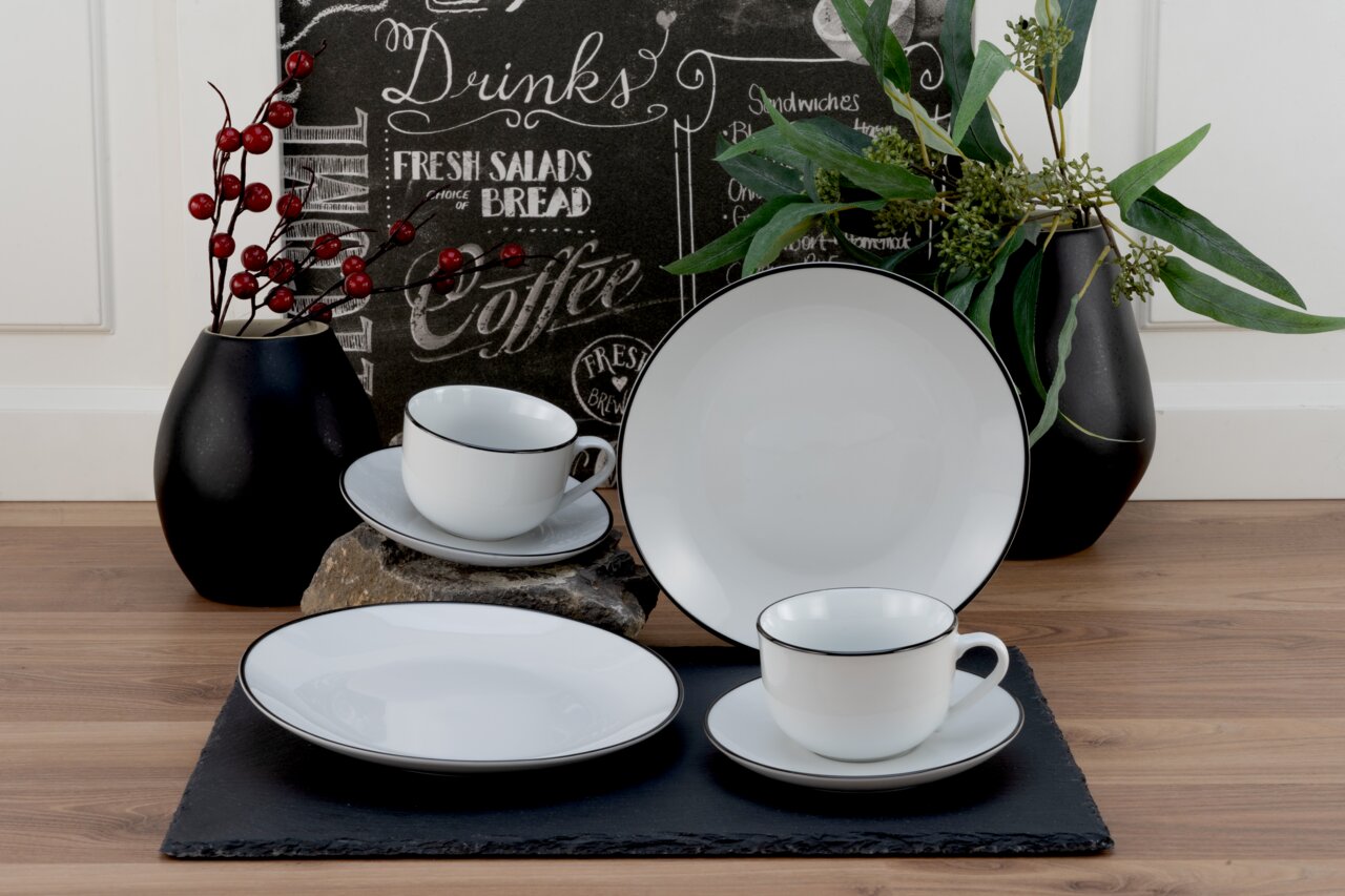 Kaffeeservice 18-teilig Luna Black L, bestehend aus weißen Tassen, Untertassen und Tellern mit schwarzem Rand, auf einem Tisch mit dekorativen Vasen und Pflanzen, seitliche Perspektive.