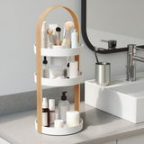 Dreistöckiger Kosmetik-Organizer mit Holzgriff, gefüllt mit verschiedenen Kosmetikartikeln, auf einer Badezimmerablage neben einem Waschbecken, aus seitlicher Perspektive.